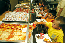 Five-year old Noah Dent (yellow shirt) grabs a slice of pizza during the last week of school at Peachland Elementary. His classmate Roberto Solano ponders his lunch choices.